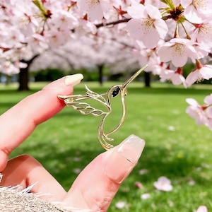 Könnte beinhalten: Goldfarbene Kolibri-Brosche mit schwarzen Akzenten. Die Brosche wird vor einem Hintergrund aus rosa Kirschblüten und grünem Gras gehalten. Der Kolibri wird im Flug mit ausgebreiteten Flügeln dargestellt.