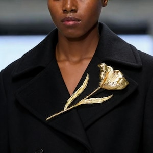 May include: A gold tulip brooch pinned to a black coat. The brooch is a detailed representation of a tulip flower with textured petals and leaves. The coat has a classic lapel design.