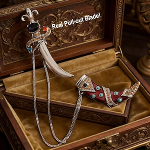 May include: A decorative dagger and a horn-shaped pendant with a pull-out blade, displayed in a wooden box. The dagger features a silver blade and is adorned with colorful gemstones. The horn pendant is red with turquoise accents.