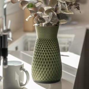 May include: A green vase with a diamond pattern, holding dried eucalyptus branches. The vase has a textured surface and a cylindrical shape, with a slightly wider top. A white mug is in the foreground.