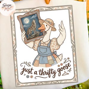 Puede incluir: Camiseta blanca con un dibujo de un ganso con sombrero y peto, sosteniendo un cuadro de un lobo aullando a la luna. El texto "just a thrifty goose" está debajo, dentro de un marco floral.