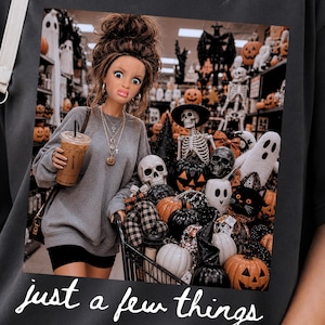 May include: A dark gray t-shirt featuring a graphic print of a doll with a high bun hairstyle holding an iced coffee, standing in a store aisle filled with Halloween decorations including pumpkins, skulls, and ghosts, with the text just a few things.
