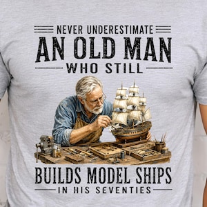 May include: A light gray t-shirt featuring a graphic of a person with gray hair and glasses wearing a blue shirt and brown apron, working on a model ship on a wooden workbench. The text reads: NEVER UNDERESTIMATE AN OLD MAN WHO STILL BUILDS MODEL SHIPS IN HIS SEVENTIES.