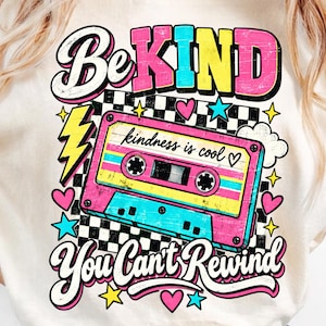 May include: A cream-colored t-shirt featuring a graphic print of a pink and blue cassette tape over a black and white checkerboard pattern. The text reads "BE KIND" at the top, "kindness is cool" on the tape, and "You Can't Rewind" at the bottom.