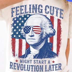 May include: A cream-colored t-shirt featuring a graphic of George Washington wearing American flag-patterned sunglasses. The design includes the text FEELING CUTE at the top and MIGHT START A REVOLUTION LATER at the bottom, set against a distressed flag.
