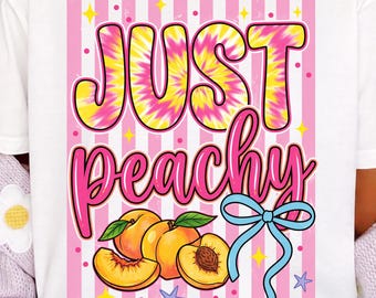 May include: White t-shirt with the text "JUST peachy" in tie-dye and pink lettering. The design includes peaches, a blue bow, and pink and white striped background. The shirt has a round neck and short sleeves.