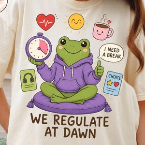 May include: A cream-colored t-shirt featuring a graphic of a green frog in a purple hoodie sitting on a purple cushion. The frog holds a clock and gives a thumbs up. Surrounding icons include a heart, sun, coffee mug, headphones, and text reading "I NEED A BREAK" and "WE REGULATE AT DAWN".