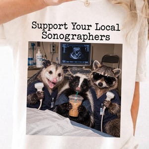 Może przedstawiać: Kremowy t-shirt z napisem "Support Your Local Sonographers" nad grafiką przedstawiającą oposa, skunksa i szopa pracza w strojach medycznych. Zwierzęta trzymają sondy ultrasonograficzne oraz kubek z kawą w gabinecie lekarskim.
