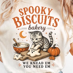 May include: A cream-coloured t-shirt featuring a graphic of a tabby cat kneading ghost-shaped dough on a wooden board. The design includes a pumpkin, a mixing bowl, a crescent moon, and the text SPOOKY BISCUITS bakery WE KNEAD EM YOU NEED EM.
