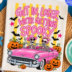 May include: A white t-shirt featuring a graphic of two skeletons in a pink convertible car. The text reads "GET IN, LOSER WE'RE GETTING SPOOKY" in pink letters. The design includes yellow vertical stripes, orange jack-o'-lanterns, and black bats.