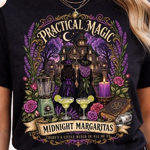 May include: A black t-shirt featuring a graphic print of two figures in witch hats standing before a large house. The design includes text reading "PRACTICAL MAGIC," "MIDNIGHT MARGARITAS," and "THERE'S A LITTLE WITCH IN ALL OF US," with illustrations of a blender, margaritas, a candle, and a book.