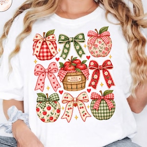Könnte beinhalten: Ein weißes T-Shirt mit einem Grafikdruck eines Holzkörbchens voller roter Äpfel, umgeben von verschiedenen dekorativen Schleifen und gemusterten Äpfeln. Auf der Vorderseite des Korbs steht der Text "APPLE PICKING".