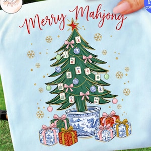 May include: A light blue t-shirt featuring a graphic of a Christmas tree in a blue and white dragon-patterned pot. The tree is decorated with mahjong tiles, pink bows, and blue ornaments, with the red text Merry Mahjong above the tree.