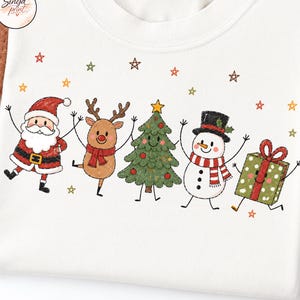 May include: A white t-shirt featuring a colourful graphic of a cartoon Santa Claus, a reindeer, a Christmas tree, a snowman, and a gift box, all arranged in a row. The design includes small scattered stars in red, orange, and green colours.