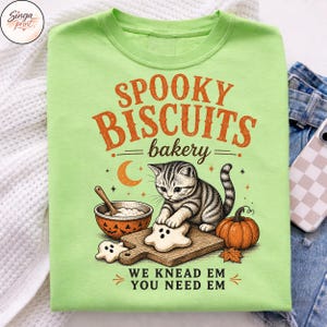Puede incluir: Una camiseta verde lima con un gr&aacute;fico de un gatito atigrado horneando galletas con forma de fantasma sobre una tabla de madera. El texto dice "SPOOKY BISCUITS bakery" y "WE KNEAD EM YOU NEED EM" junto a una calabaza y un cuenco.