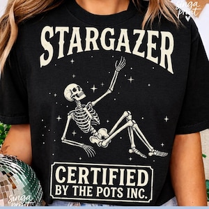 May include: Black t-shirt with the word "STARGAZER" in large white letters above a skeleton illustration. The skeleton is reaching up towards stars. Below the skeleton is the text "CERTIFIED BY THE POTS INC."