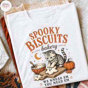 Puede incluir: Una camiseta blanca con un gr&aacute;fico de un gatito atigrado horneando galletas con forma de fantasma. El texto dice "SPOOKY BISCUITS bakery" y "WE KNEAD EM YOU NEED EM". El dise&ntilde;o incluye una luna creciente naranja, una calabaza y un taz&oacute;n.