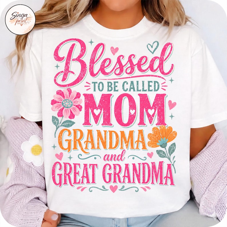May include: White t-shirt with a colourful graphic design. The design features the words "Blessed to be called MOM, GRANDMA, and GREAT GRANDMA" in pink and orange, with floral and heart accents.