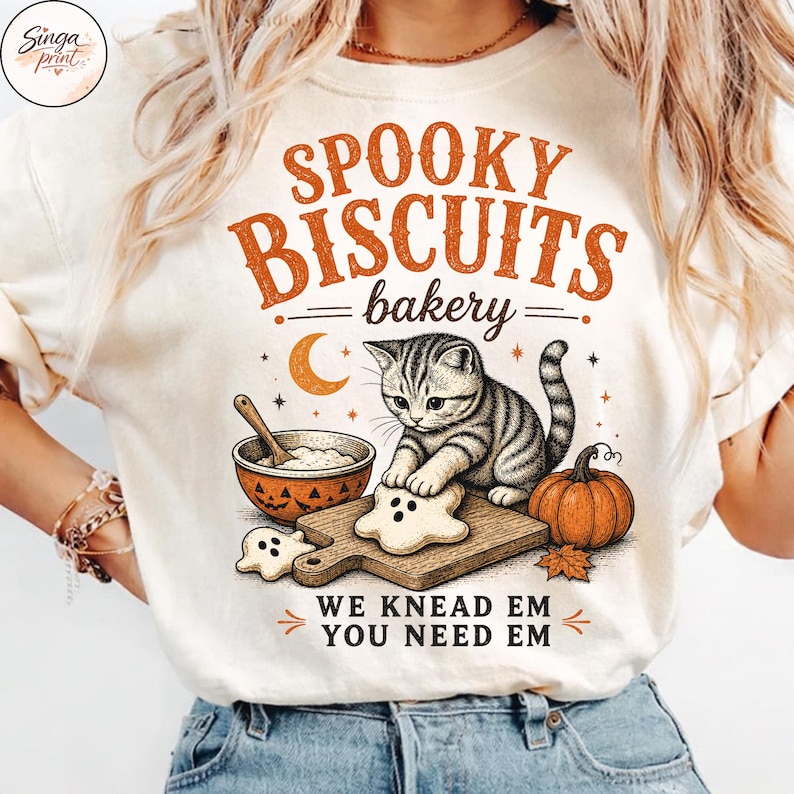 Puede incluir: Una camiseta color crema con el gr&aacute;fico de un gato atigrado amasando masa con forma de fantasma sobre una tabla de madera. El dise&ntilde;o incluye una calabaza, un cuenco, una luna creciente y el texto SPOOKY BISCUITS bakery WE KNEAD EM YOU NEED EM.