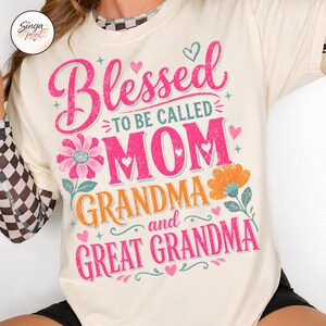 May include: Cream-coloured t-shirt with the words "Blessed to be called MOM, GRANDMA, and GREAT GRANDMA" in pink and orange lettering. Floral and heart graphics accent the text. The shirt has short sleeves.