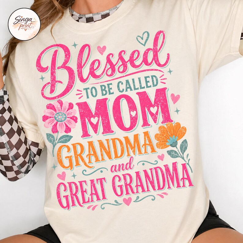 May include: Cream-coloured t-shirt with the words "Blessed to be called MOM, GRANDMA, and GREAT GRANDMA" in pink and orange lettering. Floral and heart graphics accent the text. The shirt has short sleeves.