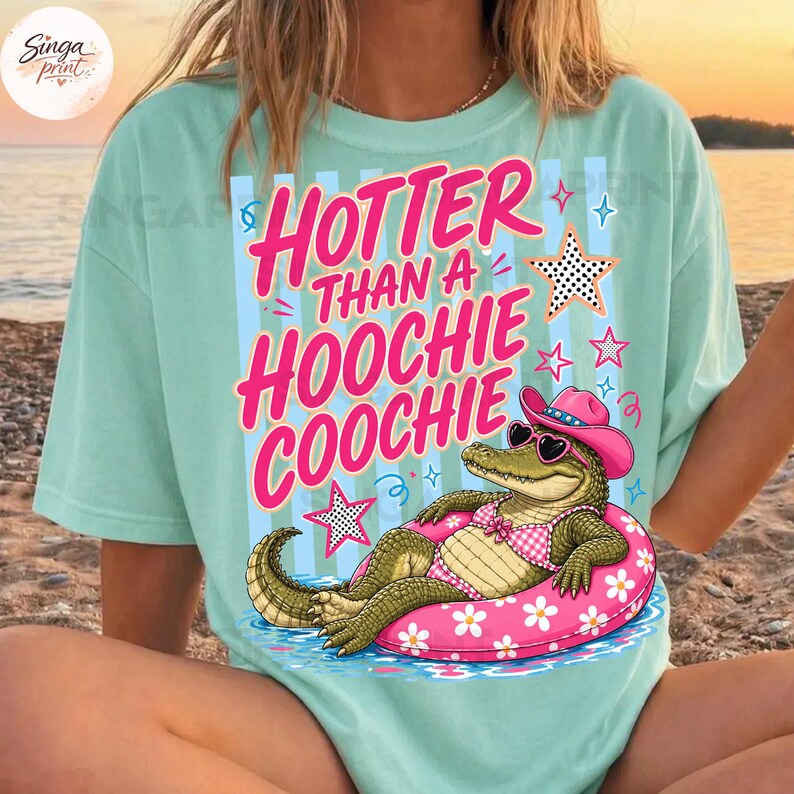 May include: A light teal t-shirt with the text "Hoter Than a Hoochie Coochie" in pink. The graphic features an alligator wearing a pink cowboy hat and sunglasses, floating in a pink inner tube. Stars and sparkles surround the alligator.