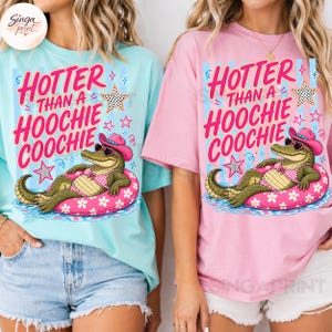 May include: Two t-shirts, one light blue and one pink, each featuring a graphic of an alligator in a pink floatie, wearing a cowboy hat and sunglasses. The text on the shirts reads "HOTTER THAN A HOOCHIE COOCHIE".