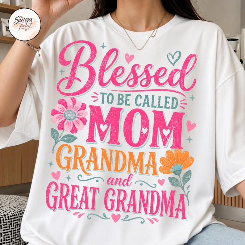 May include: White t-shirt featuring the text "Blessed to be called Mom, Grandma, and Great Grandma" in pink and orange. The design includes floral and heart motifs, ideal for celebrating mothers.