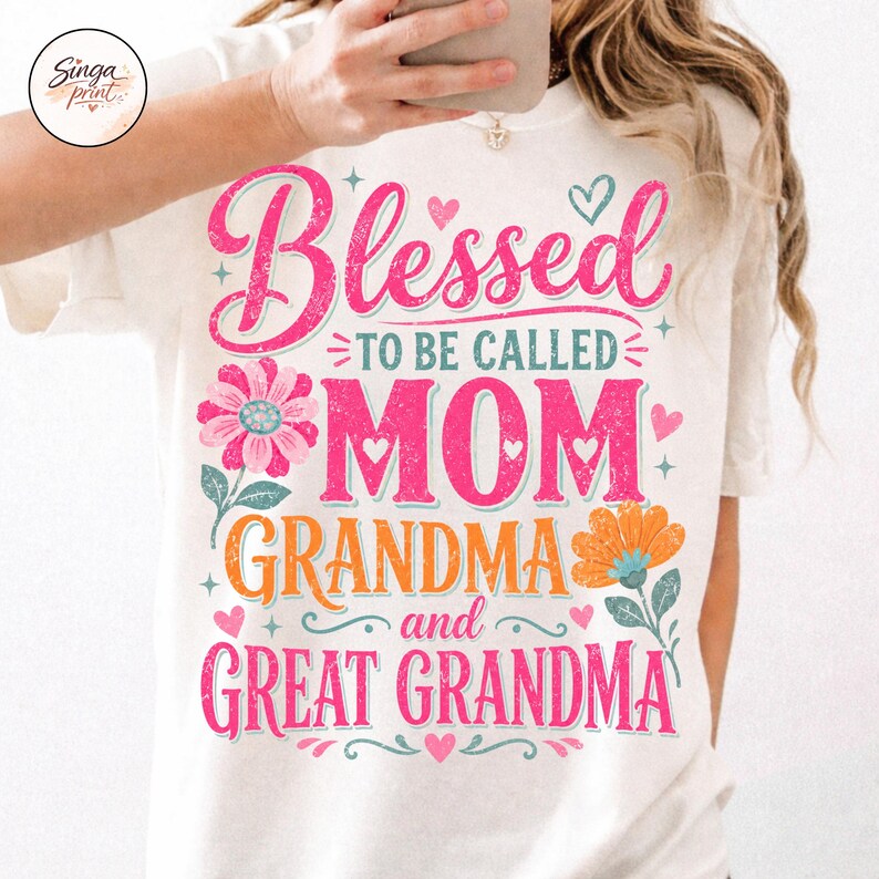 May include: A cream-coloured t-shirt featuring the text "Blessed to be called Mom, Grandma, and Great Grandma" in pink and orange. The design includes floral and heart graphics, creating a cheerful and celebratory look.