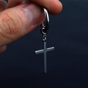 Puede incluir: Un pendiente de aro de metal plateado con un colgante de cruz. El pendiente tiene un acabado pulido y liso con un diseño sencillo y limpio. La cruz está unida al aro circular mediante una pequeña anilla de metal, creando un estilo colgante.