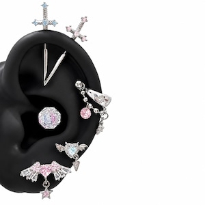May include: A collection of silver-toned ear piercings on a black ear model, featuring cross designs with blue and pink stones, a faceted circular stud, a dangling charm piece, and heart-shaped motifs with wings and hanging star accents.