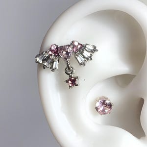 May include: A silver-toned ear cuff and stud earring set. The ear cuff features pink and clear crystals in a fan design with a dangling star. The stud earring is a single pink crystal. Jewelry.