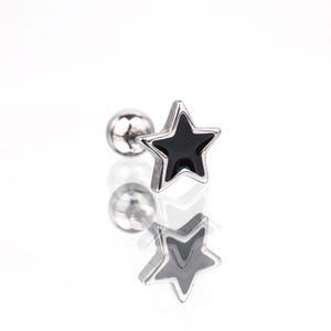 May include: A silver-toned metal stud earring featuring a five-pointed star shape with a glossy black enamel inlay. The earring includes a round ball backing and a straight post, designed for piercing, with a reflective surface visible below.
