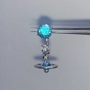 Könnte beinhalten: Ein silberfarbener Bauchnabelpiercing mit einem großen, runden, leuchtend blauen facettierten Edelstein an der Oberseite. Darunter hängt ein Anhänger, bestehend aus einem kleinen klaren Edelstein und einer blauen, planetenförmigen Perle mit Ring.