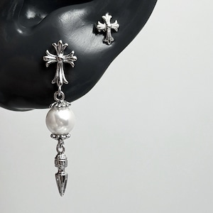 May include: A silver-toned earring set featuring a small cross stud and a dangling earring. The dangling piece consists of a silver cross top, a white spherical pearl bead, and a pointed silver charm at the base, all with metallic finishes.