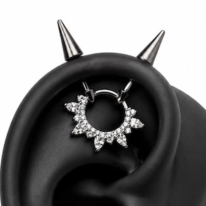May include: A dark metal septum ring featuring a circular band adorned with a sunburst of clear, faceted crystals. The jewelry is displayed on a black ear model that includes two pointed, cone-shaped spikes protruding from the top edge.