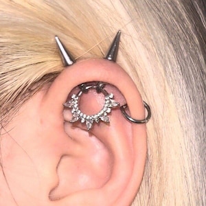 May include: Close-up of an ear adorned with multiple piercings. A silver, sunburst-shaped earring with clear stones is centered. Two silver spikes are positioned above the ear, and a simple hoop earring is visible.