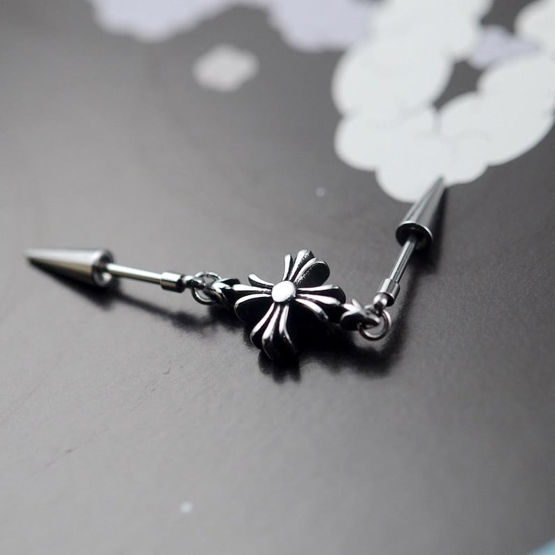May include: A silver-colored earring with a cross-shaped charm and a pointed end. The earring is connected by a chain and is set against a dark background. The cross has a detailed, floral design.