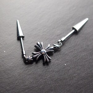 May include: A silver-colored earring with a cross-shaped charm and two conical ends. The earring is connected by a chain and is designed to be worn through multiple piercings. The jewelry is made of a shiny metal.