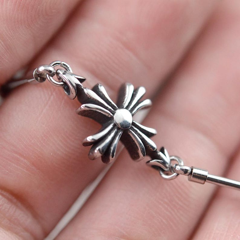 May include: Close-up of a silver-colored cross pendant. The cross features a detailed, ornate design with a central circular element and pointed edges. The pendant is attached to a thin chain.
