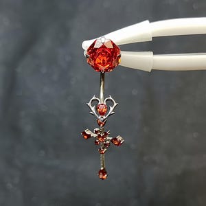 May include: A silver-toned navel ring featuring a large, faceted, round orange-red gemstone at the top. Below the curved barbell, a heart-shaped metal frame holds a small orange-red stone, followed by a cross-shaped pendant set with multiple orange-red stones.