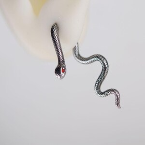 K&ouml;nnte beinhalten: Ein silberfarbener Schlangenohrring mit texturiertem Schuppenmuster und einem kleinen, leuchtend roten Stein als Auge. Das Design umschlie&szlig;t das Ohrl&auml;ppchen, wobei der Kopf vorne und der lange, gewundene Schwanz hinter dem Ohr positioniert ist.