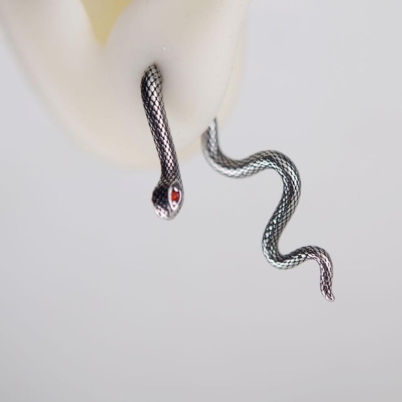 K&ouml;nnte beinhalten: Ein silberfarbener Schlangenohrring mit texturiertem Schuppenmuster und einem kleinen, leuchtend roten Stein als Auge. Das Design umschlie&szlig;t das Ohrl&auml;ppchen, wobei der Kopf vorne und der lange, gewundene Schwanz hinter dem Ohr positioniert ist.