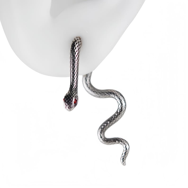 K&ouml;nnte beinhalten: Ein silberfarbener Schlangenohrring mit texturiertem Schuppenmuster und kleinen roten Augen. Das Design besteht aus einem geraden Kopfbereich, der durch das Ohrl&auml;ppchen f&uuml;hrt, und einem langen, gewundenen Schwanz, der unter dem Ohr h&auml;ngt.