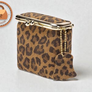 Op de afbeelding: Een rechthoekige clutch met een luipaardprint in bruin en zwart. Het heeft een goudkleurige sluiting en een bijpassende bedel die aan een ketting hangt. Het merklogo "VIBEPOUCH" staat in een cirkel linksboven.