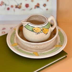May include: A cream-colored ceramic creamer, bowl, and plate set. The set features a green band with yellow chicks. The creamer and bowl are stacked on the plate. The set is on top of a green book.