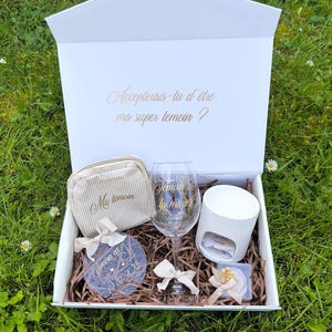 Peut inclure: Une boîte cadeau blanche contenant une pochette beige rayée, un verre à vin avec texte doré, un chauffe-bougie blanc côtelé, un ornement circulaire en acrylique avec texte doré et une petite boîte cadeau avec ruban, sur un lit de papier brun.