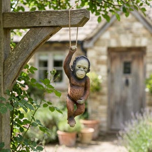 May include: A brown and tan resin orangutan statue hanging from a wooden beam by a rope. The statue has a detailed face and is suspended in mid-air. The background includes a house and greenery.