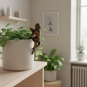 Puede incluir: Una figura de hada de bronce decora una maceta de cerámica blanca llena de plantas verdes. El hada tiene alas detalladas y está posada como si estuviera trepando por la maceta. La maceta está sobre una superficie de madera clara.