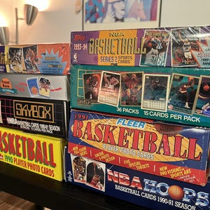 Puede incluir: Una pila de cajas de cartas de baloncesto de la NBA vintage. Las cajas presentan colores y gráficos vibrantes, con textos como "NBA Basketball" y "Fleer". Las cajas son de varias temporadas, incluyendo 1990-91 y 1993-94.